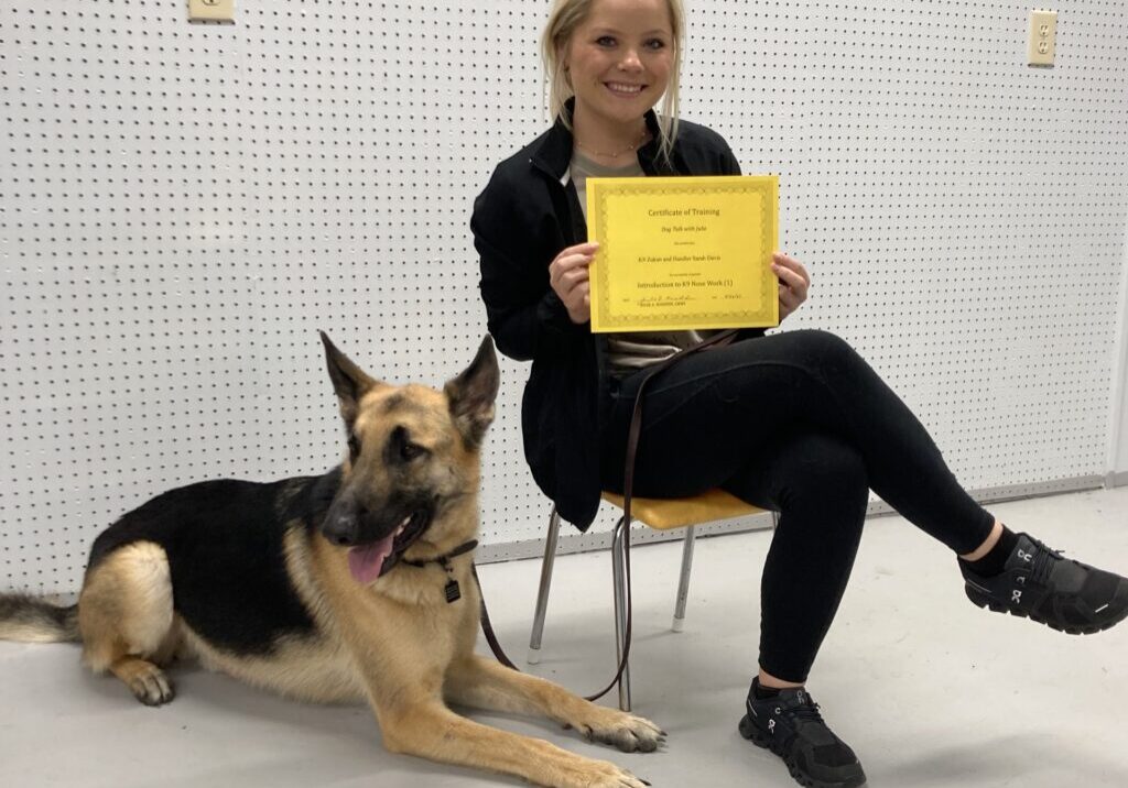 A woman holding a yellow certificate while sitting on a chair and has a dog beside her